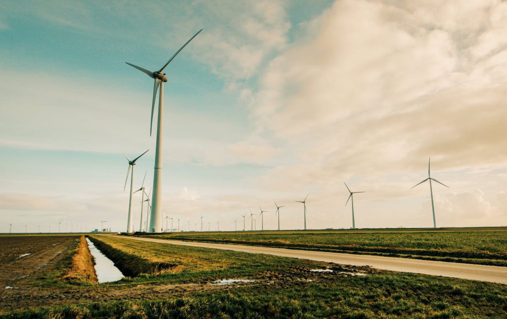 A imagem mostra algumas turbinas de energia eólica, sob um gramado, em ambiente externo, durante o dia.
