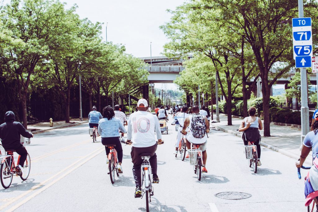 Imagem de várias pessoas de costas andando de bicicleta em uma rua arborizada, durante o dia, como exemplo de projeto de mobilidade sustentável.
