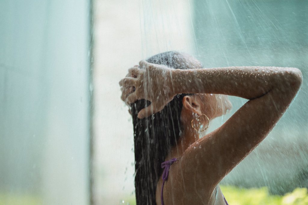 Mulher adulta, vista de lado, posiciona as duas mãos na cabeça enquanto toma banho de chuveiro.