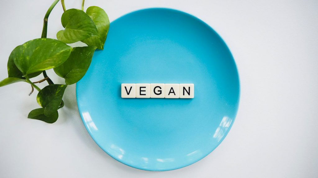  Imagem apresenta um prato de cor azul, em cima de uma superfície clara, com uma planta ao lado. No centro do prato está escrito a palavra “vegan” em inglês, que significa “vegano”.