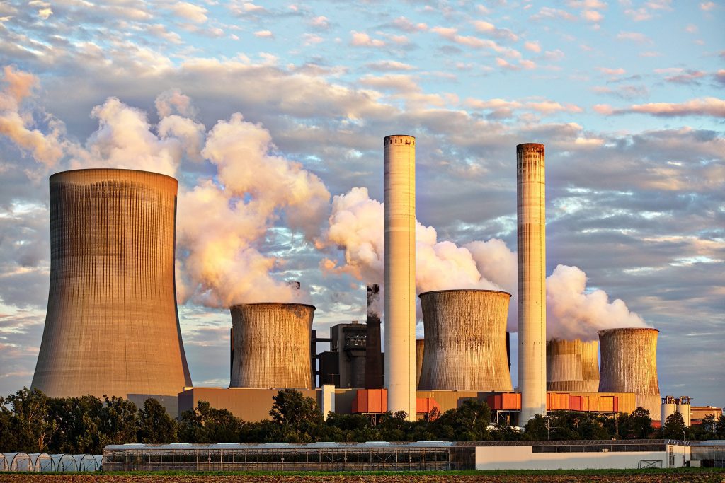 Imagem representando fontes de energia não renováveis. Uma usina nuclear é vista de dia. Há um prédio branco, com várias chaminés enormes soltando fumaça.