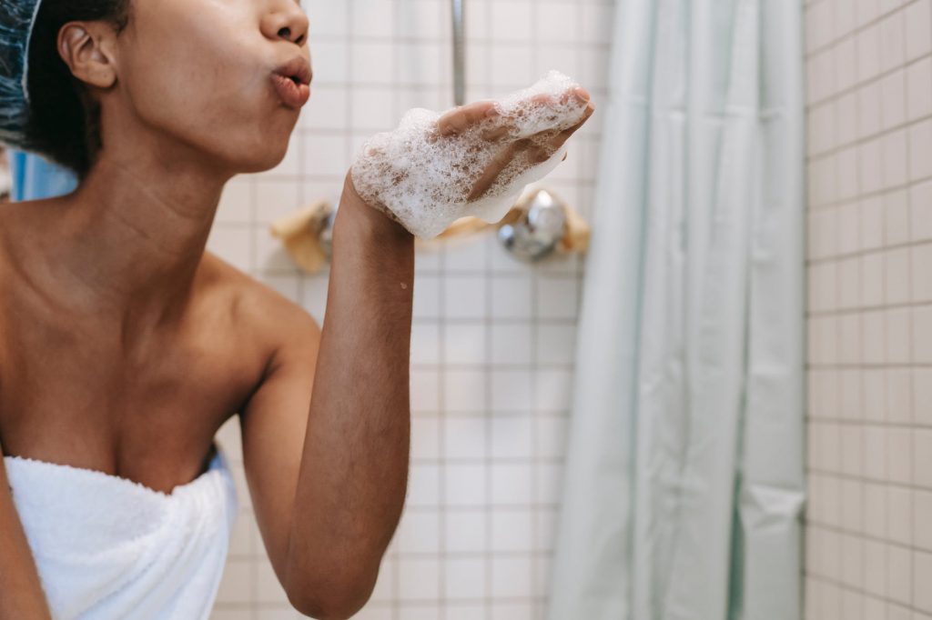 Imagem de uma mulher dentro de um banheiro, vestindo uma toalha e uma touca em seus cabelos. Ela tem espuma de banho em uma de suas mãos e faz gesto de que está soprando. Ela parece estar com o chuveiro desligado enquanto se ensaboa, uma das práticas de consumo consciente de água. 