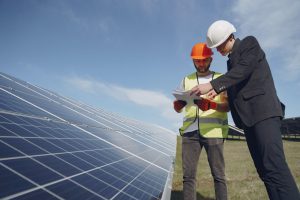 Imagem de um engenheiro e um empresário conversando enquanto observam uma folha. Eles estão em frente a um grande painel de energia solar localizado em um vasto gramado.